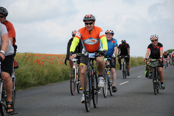 Man in an orange t-shirt cycling a bike on a road in a fundraising event