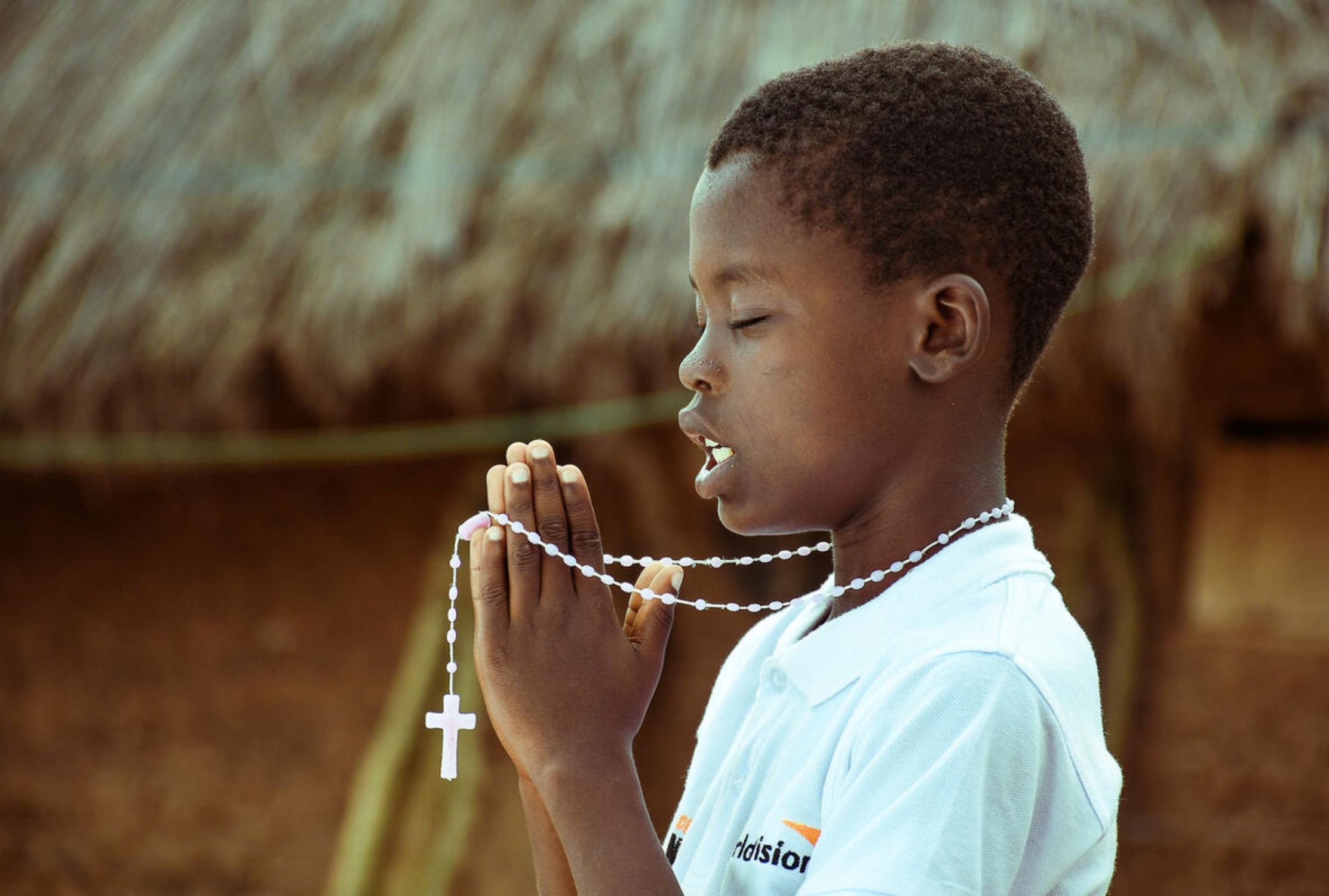 Ghanaian boy with eyes closed and hand pressed together praying, his cross necklace drapes over his fingers