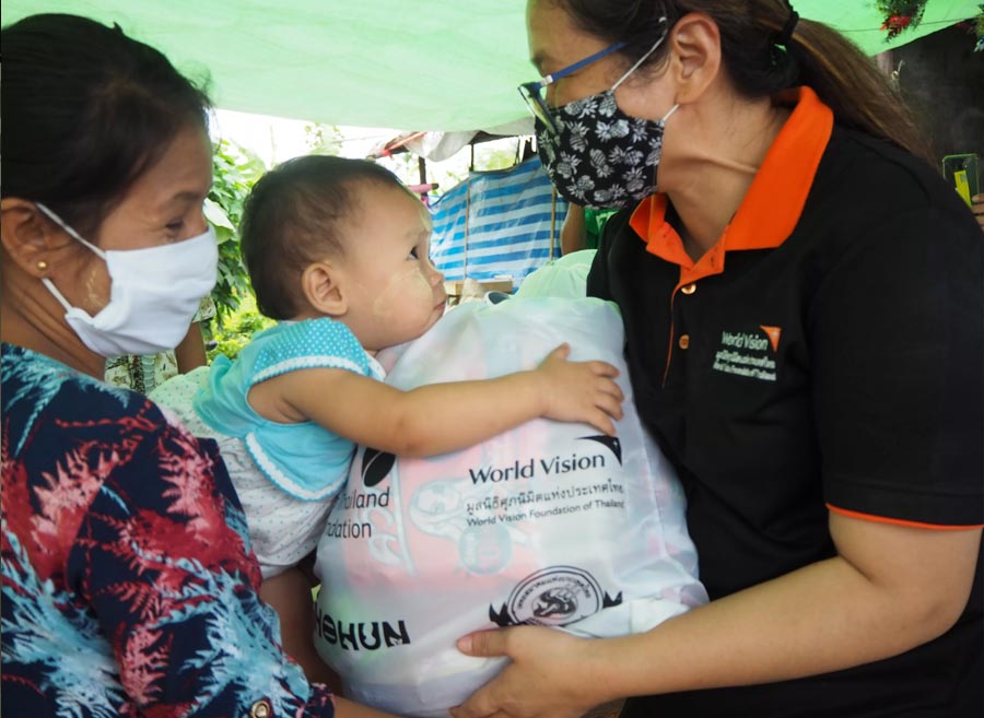 Staff member in a mask hands over a bag to support families and prevent the spread of COVID-19 in Thailand. The woman receiving the bag smiles as her child clings onto the bag