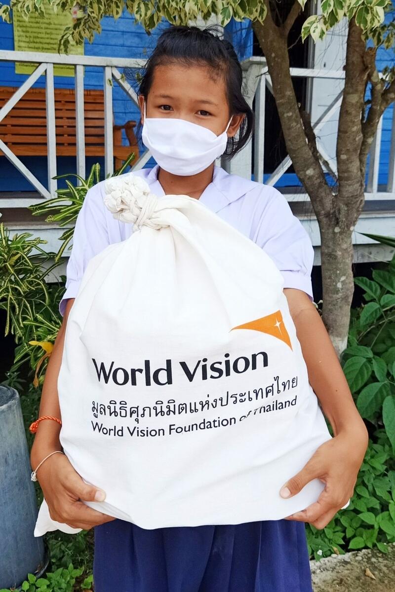 Child in Thailand wears a mask as they hold a big bag of World Vision supplies
