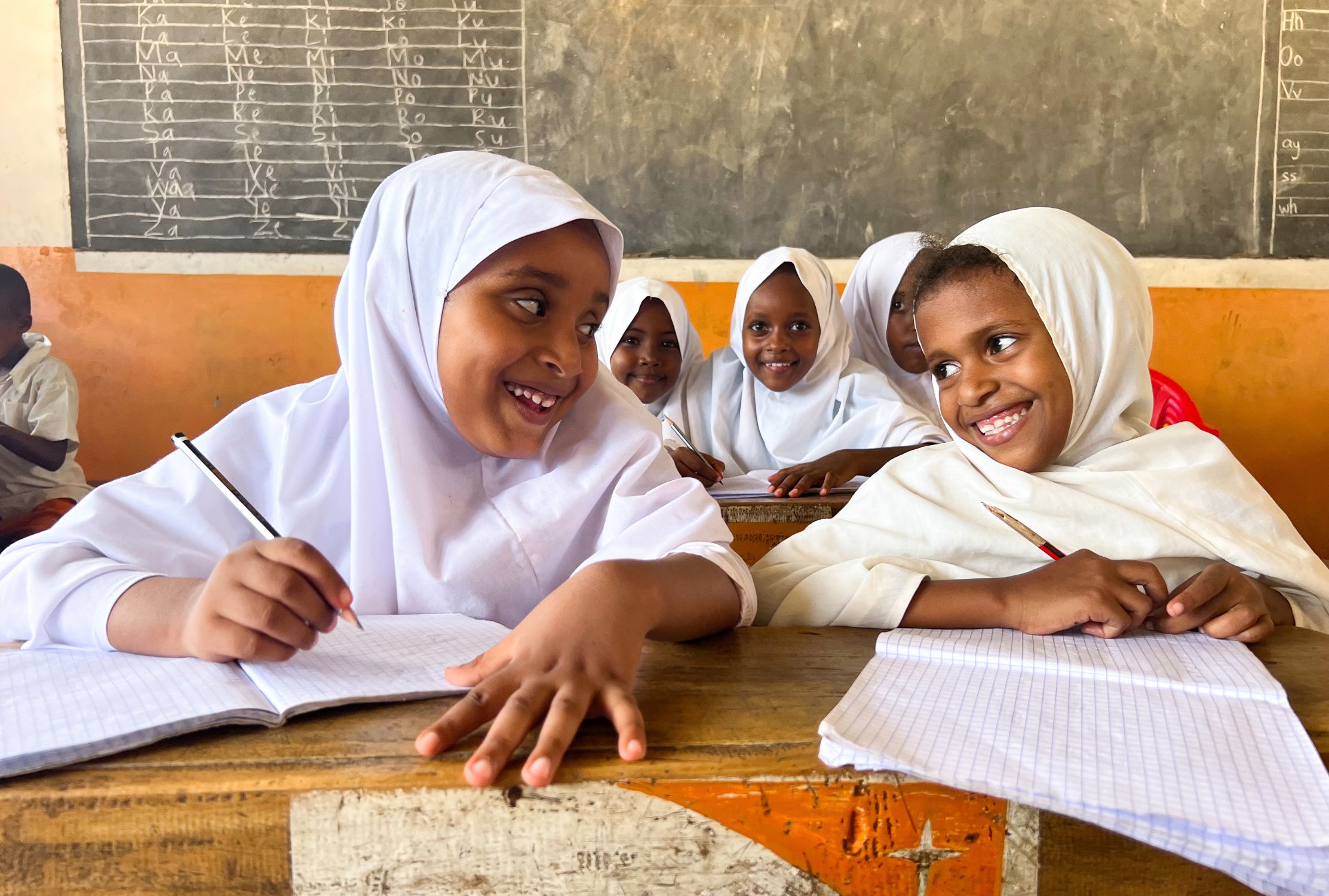 Kenyan girls smiling each other while sat at a desk with their school books. They're both wearing white hijabs