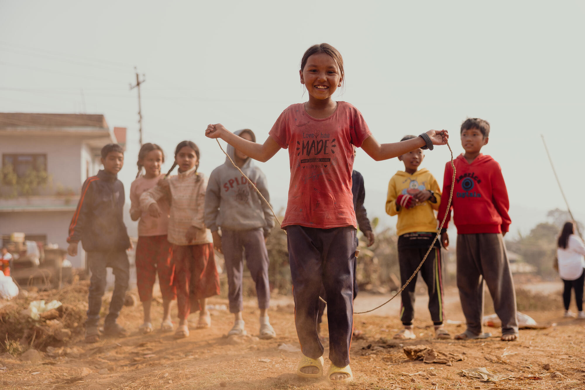 A girl skips with a rope on a dusty path in Nepal, while other children stand nearby in an outdoor community setting.