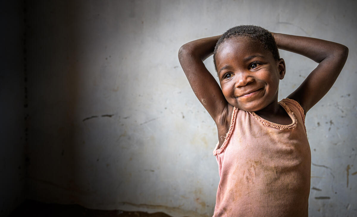 Child stands in a room in Malawi, hands clasped behind their head, smiling