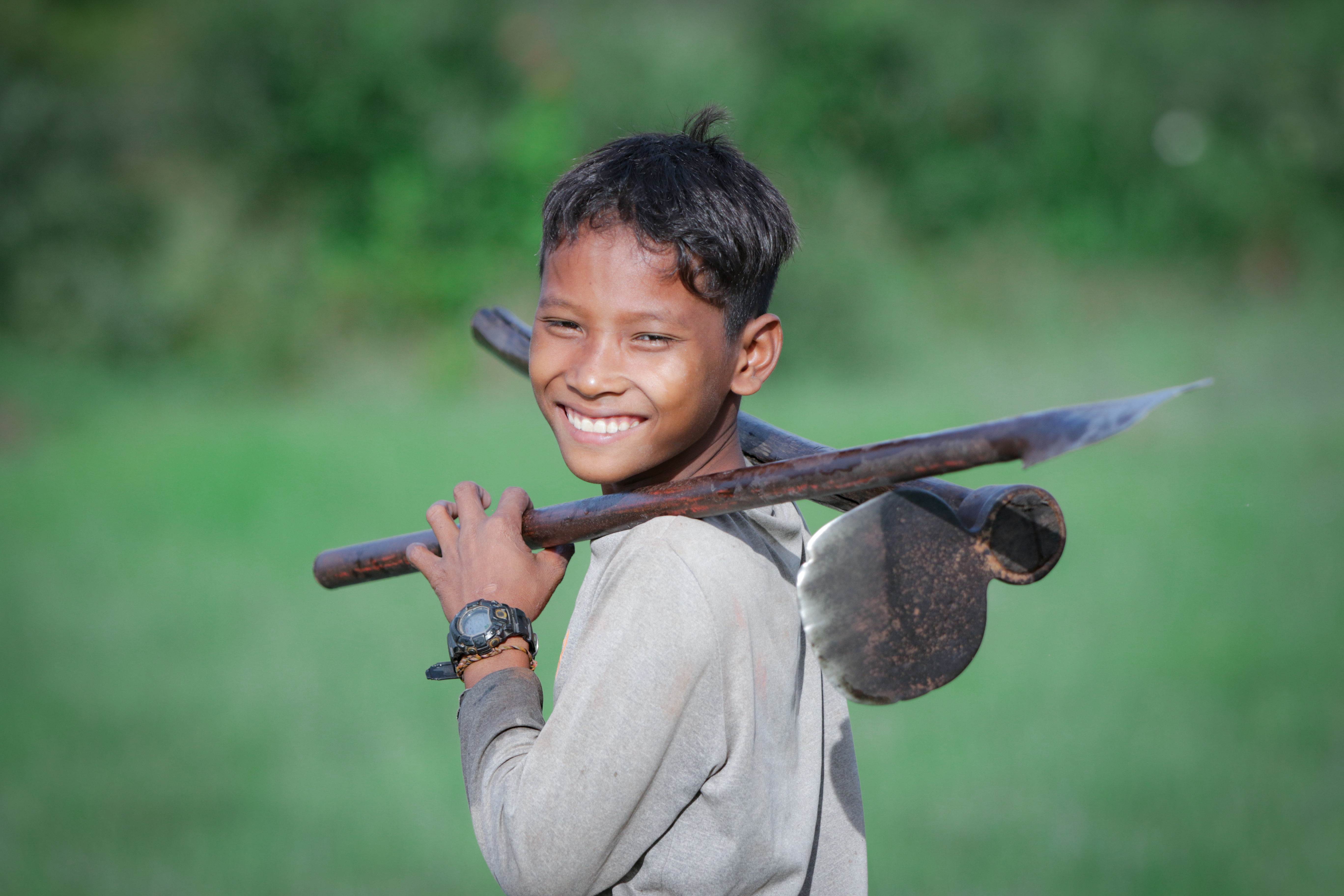 Boy in Cambodia smiles as he looks over his shoulder, standing with tools over his shoulder
