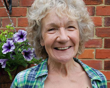 Woman in the UK smiles broadly, standing against a brick wall and in front of flowers