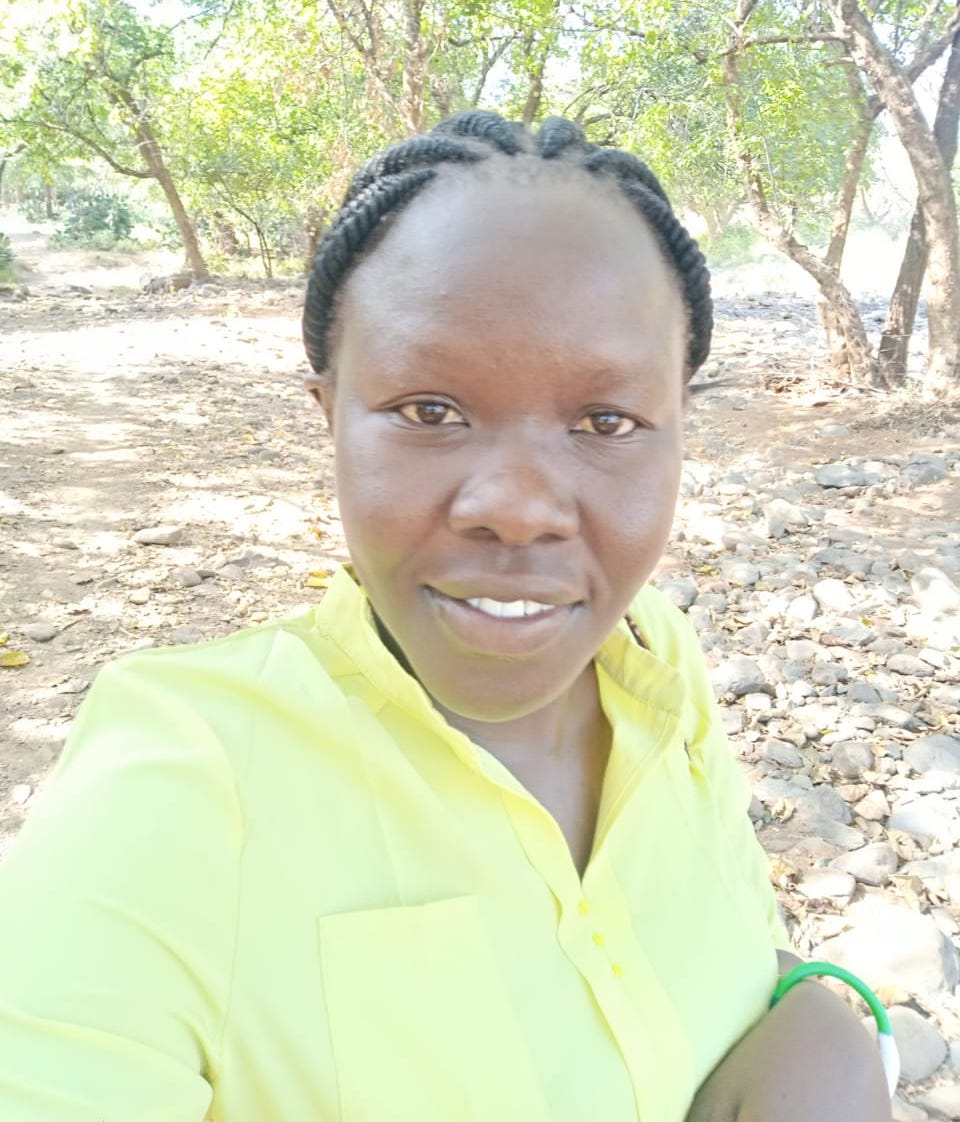 Selfie photo of young woman with braided hair and yellow top, smiling at the camera. Dusty ground and trees behind her.