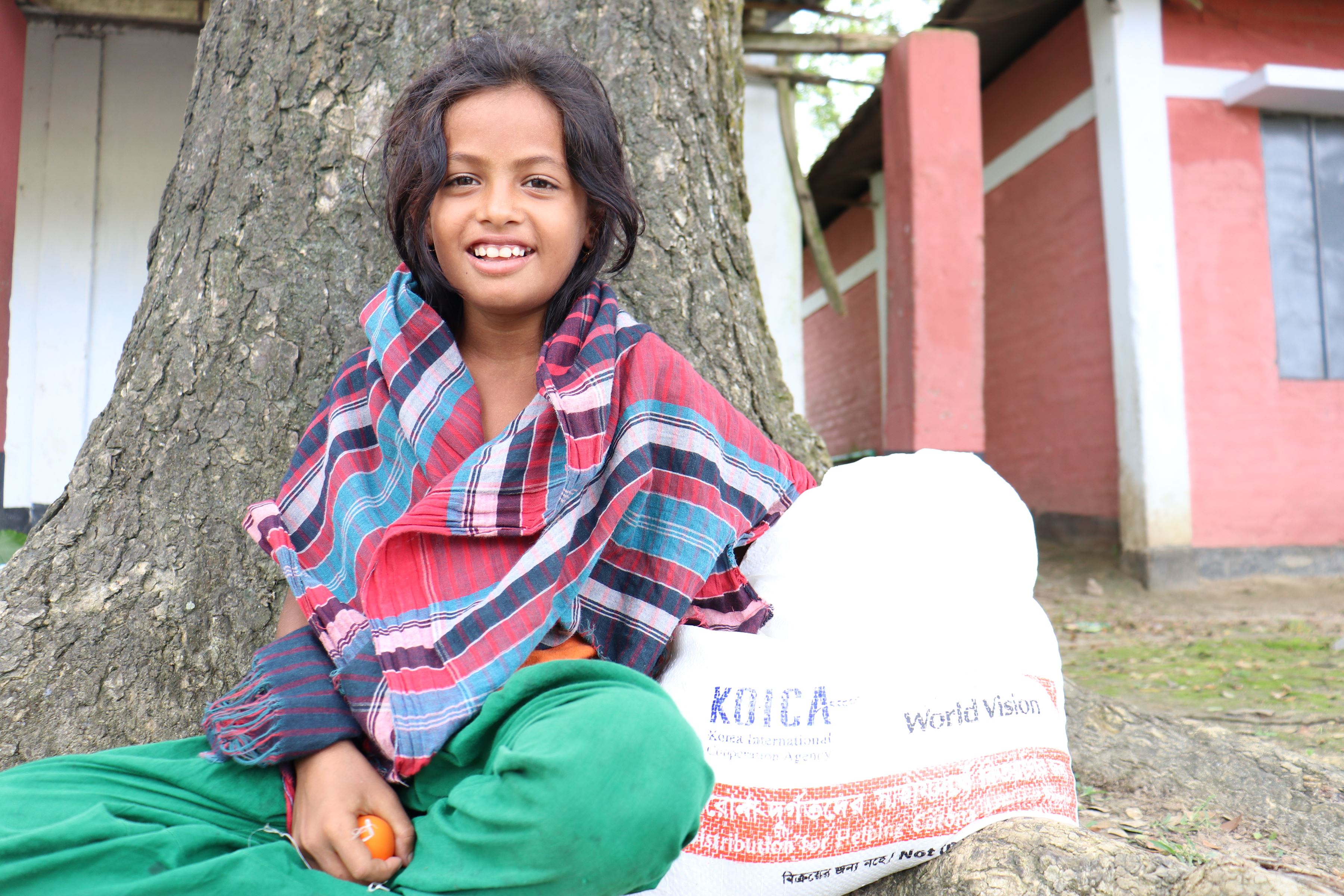 Girl sits in front of a treetrunk in Bangladesh, holding the bag of rice she's been given from a food distribution programme