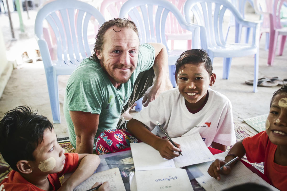 Jerome Flynn sits with young boy in Myanmar, the child is working in a workbook with a pen as Jerome smiles